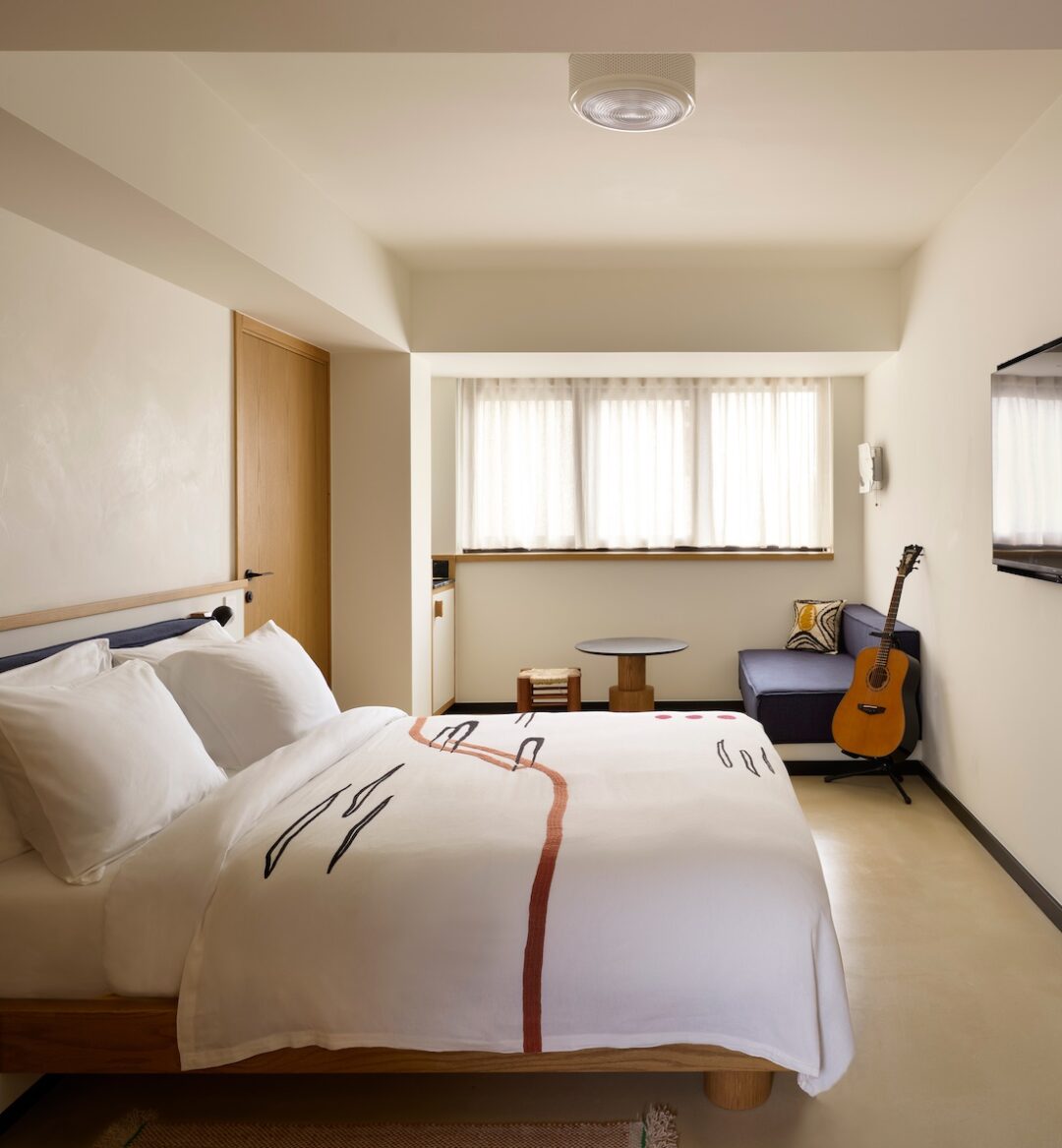 Cozy hotel room with a soft, white duvet and abstract design. A guitar leans against a blue sofa near a window. Warm tones, serene atmosphere.