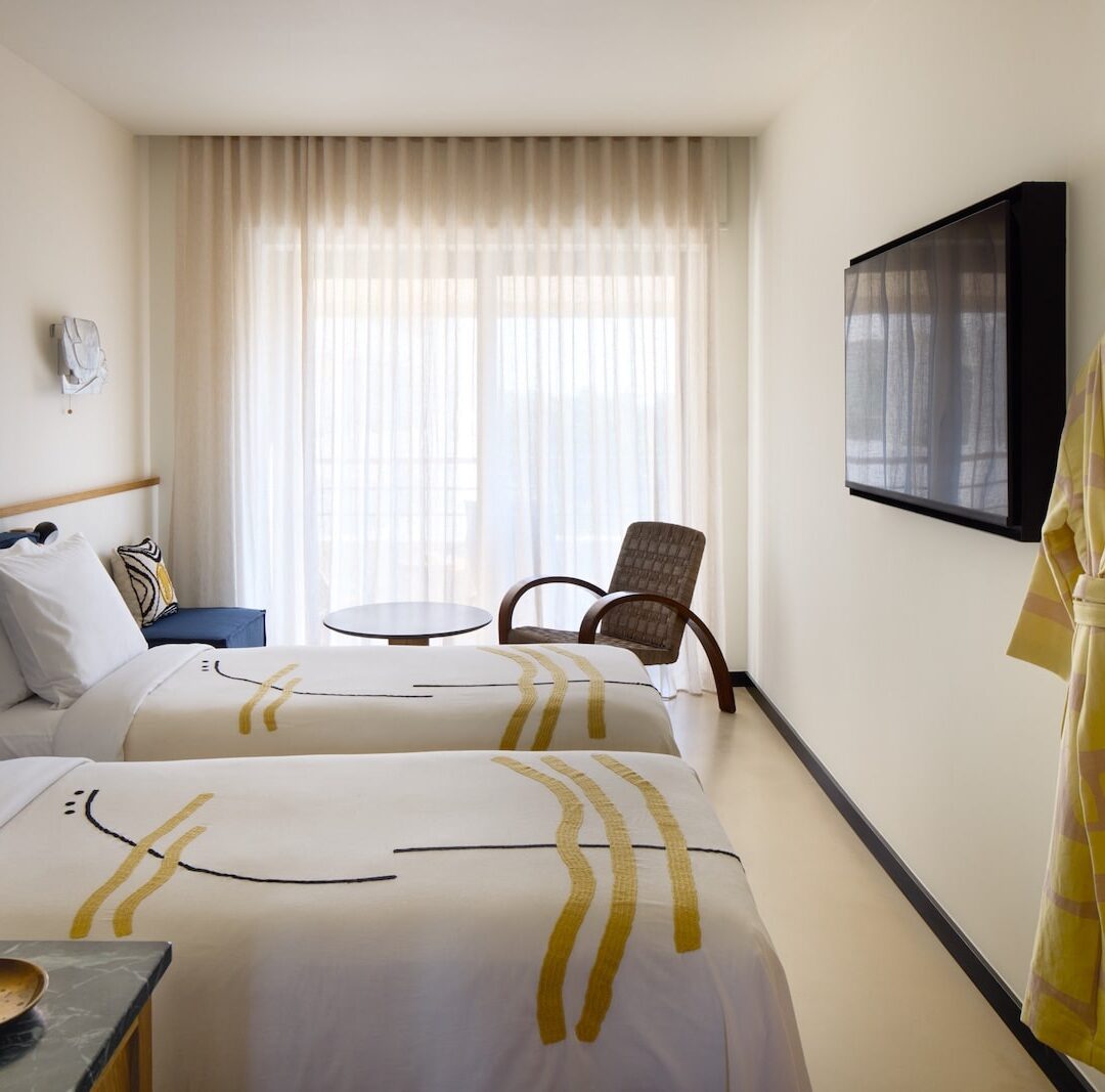 A modern hotel room with two neatly made beds featuring abstract patterned bedspreads. A large window with sheer curtains provides soft natural light. A cozy chair and small round table are near the window, and a wall-mounted TV is opposite the beds, creating a calm, inviting atmosphere.