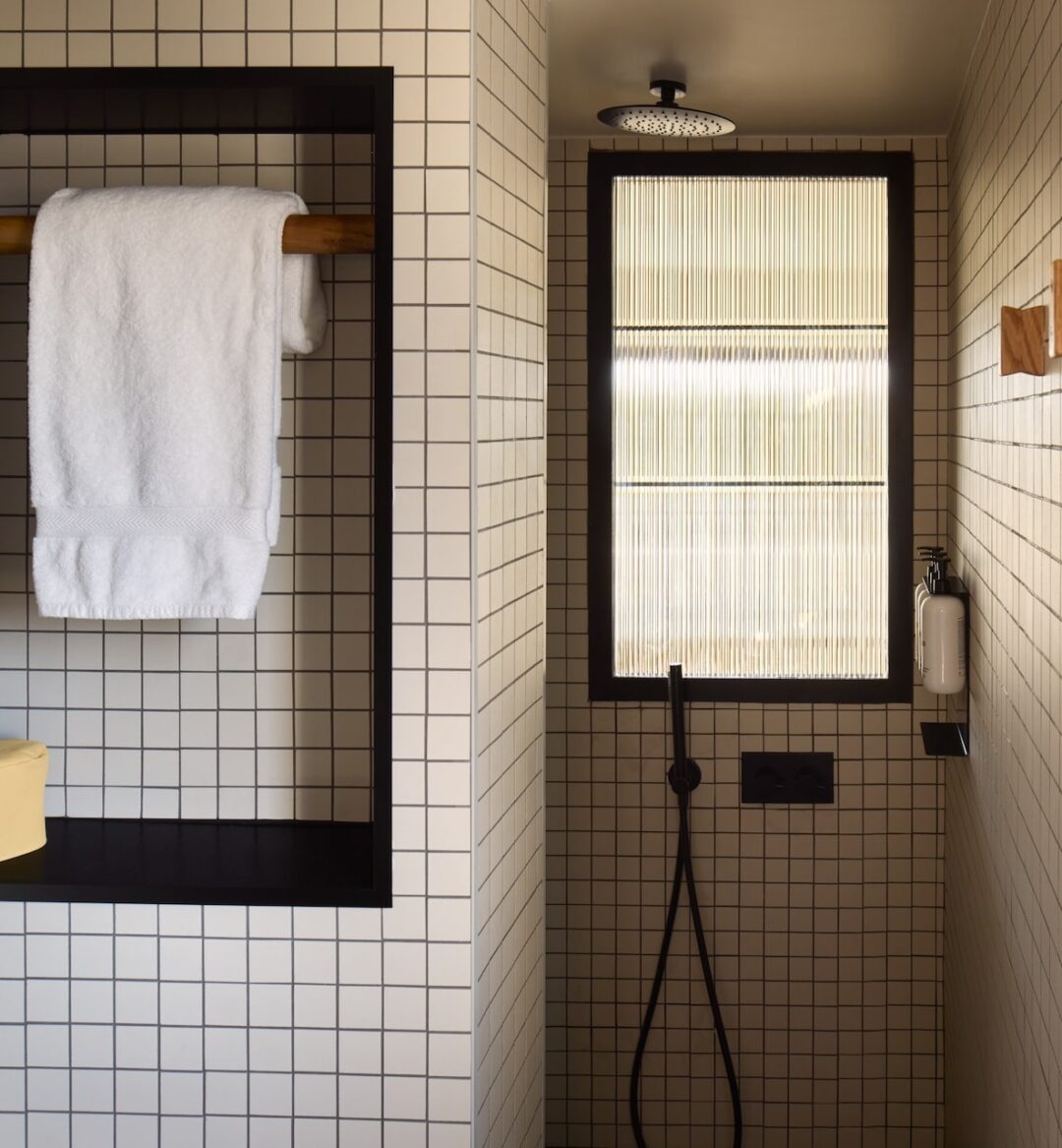 Modern bathroom with grid-patterned tiles, featuring a recessed shelf with a white towel and yellow soap. A rain shower head and a ribbed glass window add elegance.