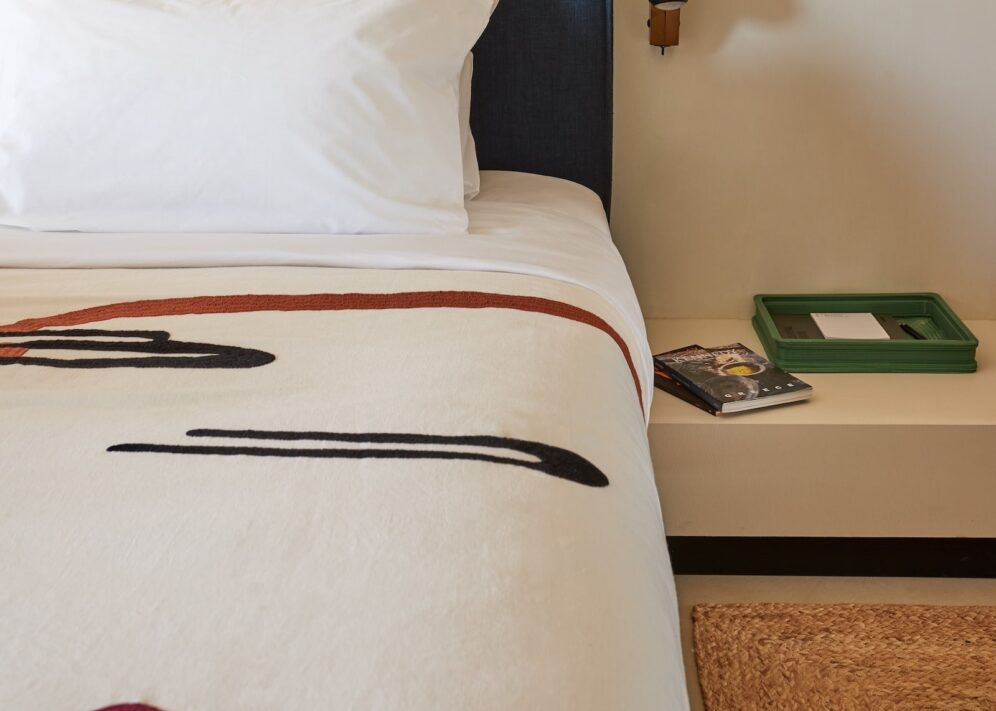 A neatly made bed with a white pillow and a blanket featuring abstract black and red lines. A nightstand holds a green tray and a book, creating a minimalist and calm atmosphere.