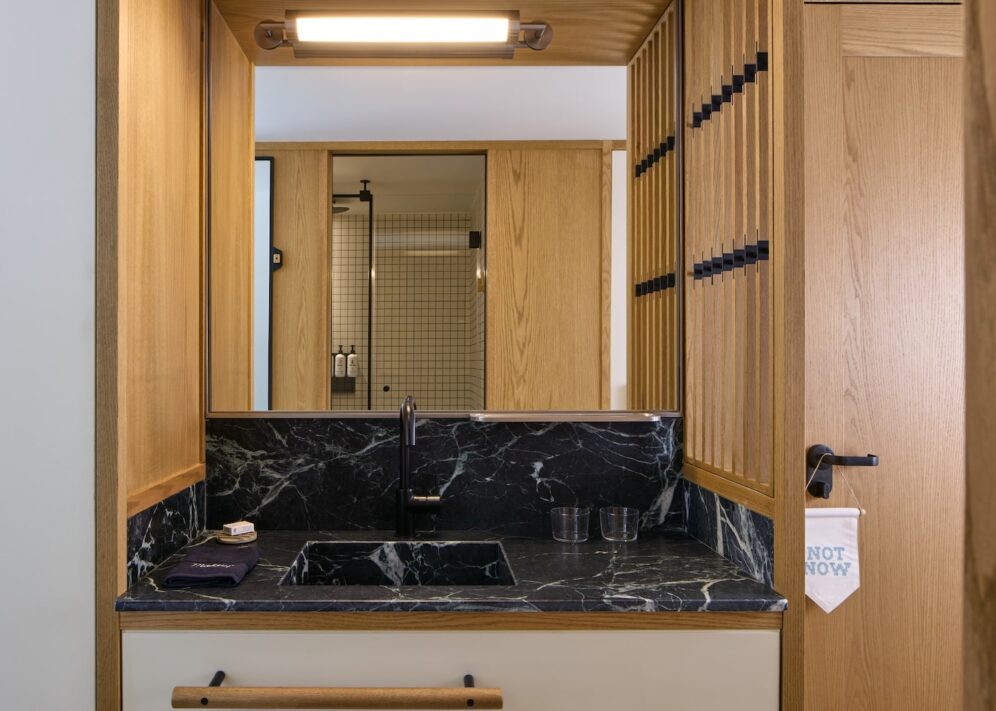 Modern bathroom with a black marble sink, wooden walls, and a rectangular mirror reflecting a shower with tiled walls. Warm, minimalist design.