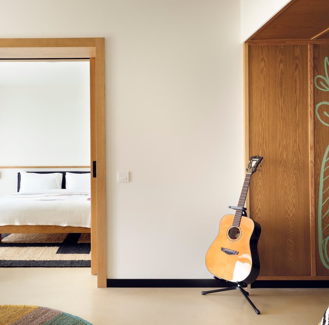 A serene room features a wooden door leading to a cozy bedroom with a white bed. A guitar stands beside a wood panel with artistic green designs.