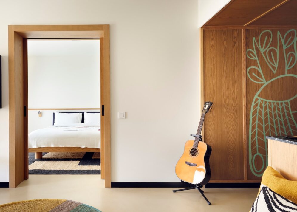 A serene room features a wooden door leading to a cozy bedroom with a white bed. A guitar stands beside a wood panel with artistic green designs.
