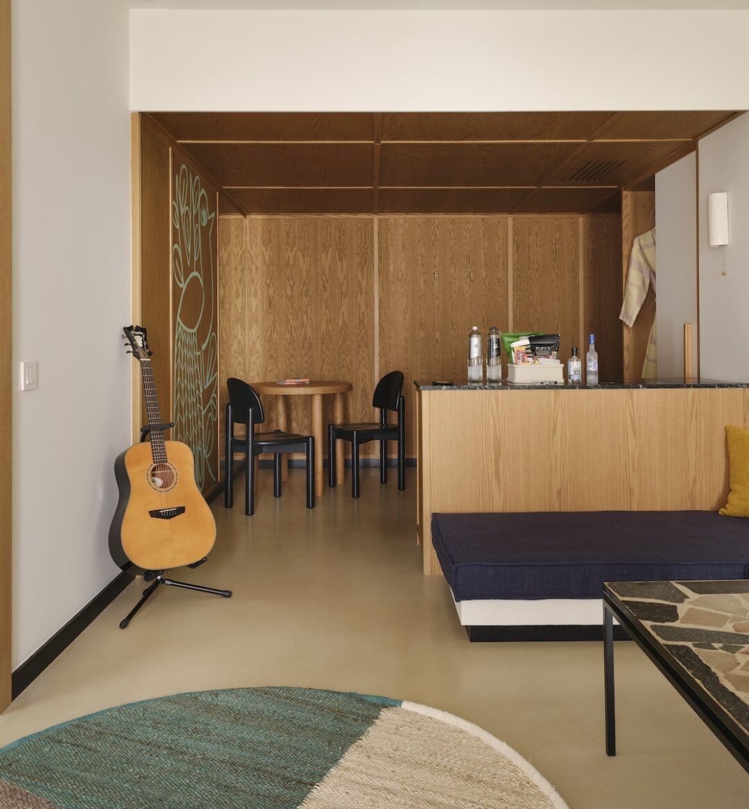 Cozy living space featuring a guitar on a stand, wood-paneled walls, blue sofa with vibrant cushions, mosaic coffee table, and dining area. Warm, inviting ambiance.