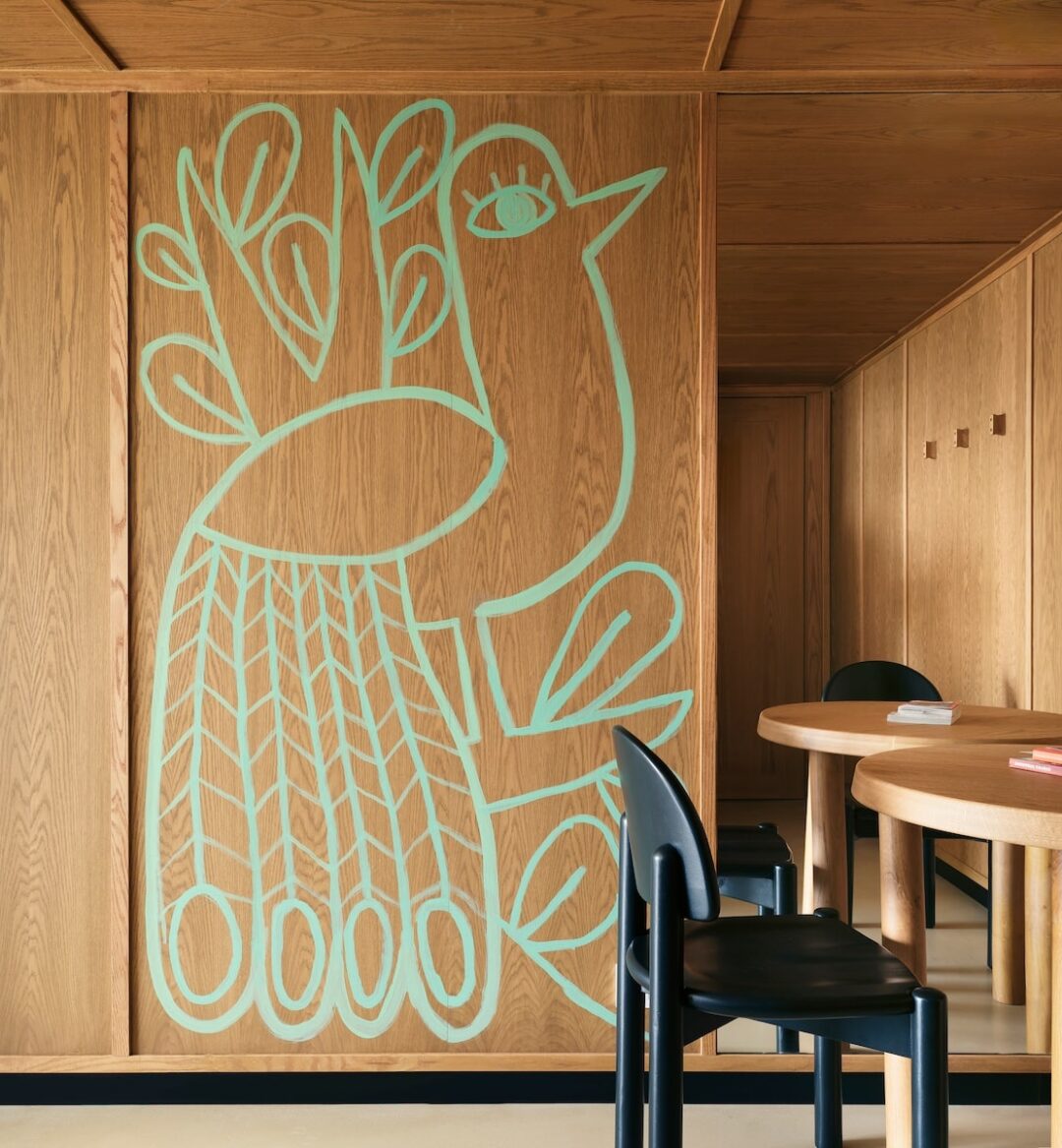 A wooden room features an abstract teal bird design on the wall. The room has a warm and cozy ambiance, with a wooden table and black chairs.