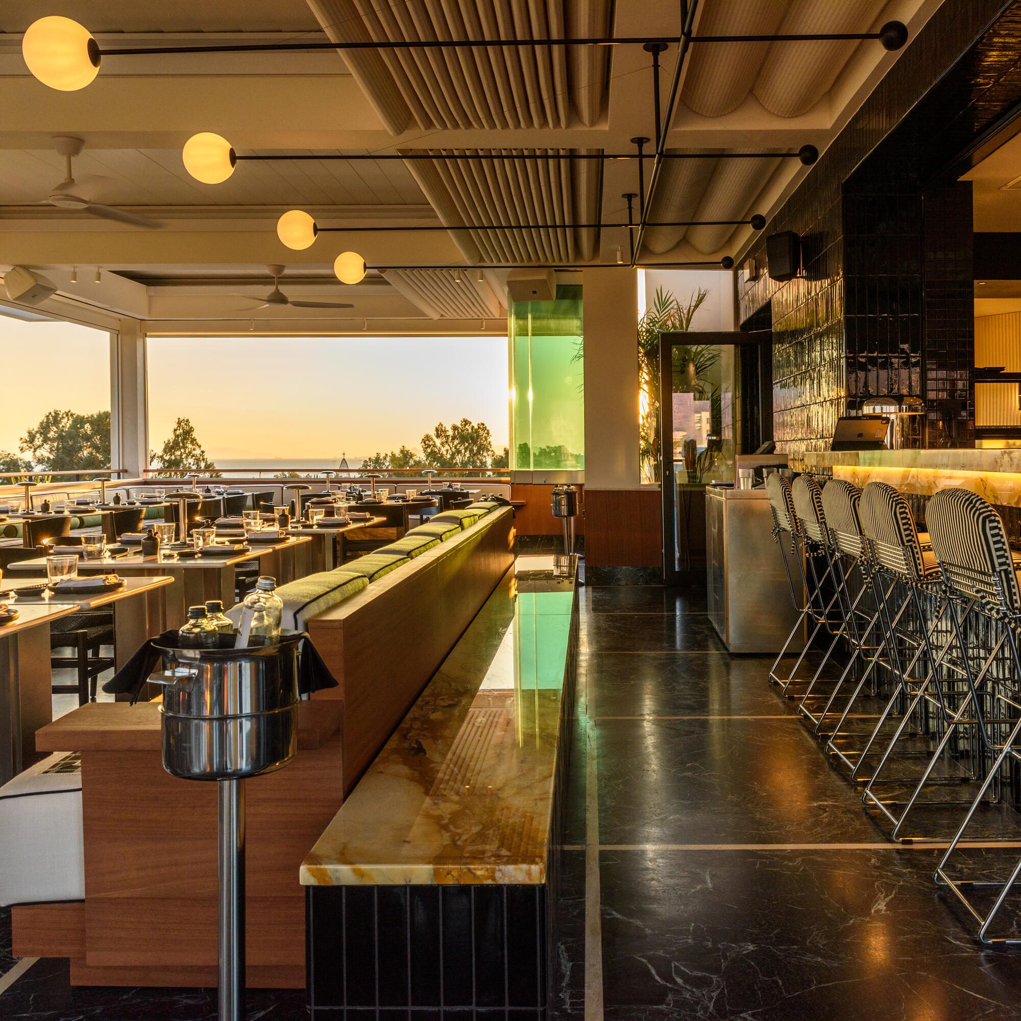 Sora at Ace Hotel Athens: a long onyx counter and dark marble floor running toward an open wall and the sea at sunset.
