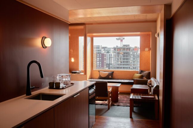 Modern hotel room with kitchenette, seating area by large window, city buildings visible outside, warm wood tones throughout.